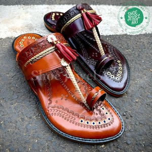 May include: Two leather sandals, one in a rich brown and the other in a deep burgundy. Both feature intricate embossed patterns, gold accents, and red tassels. The sandals have a toe loop design.