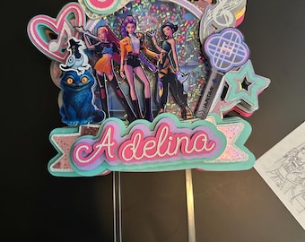 May include: A multi-layered cake topper with a holographic center, featuring a group of stylized figures and the name "Adelina". The topper includes a number "6", stars, musical notes, and a blue cat figure. It is mounted on clear plastic sticks.