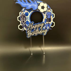 May include: A layered cake topper with a soccer theme. The design features two anime-style figures, a soccer ball, and the words "Happy Birthday." The color scheme includes shades of blue, white, and black. The topper is mounted on clear acrylic sticks.