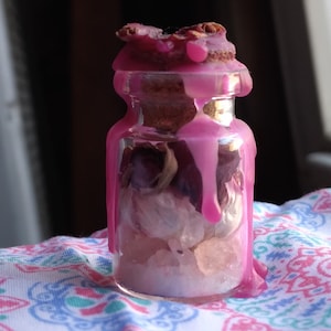 May include: A small glass jar filled with crystals and dried flowers, sealed with a cork and pink wax. The wax drips down the sides of the jar, creating a decorative effect. The jar sits on a patterned fabric.