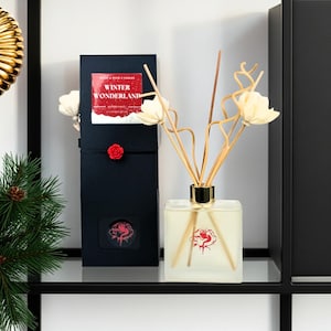 Puede incluir: Una botella difusora cuadrada de vidrio con un diseño de rosa roja, llena de líquido y cañas. Una caja negra con las palabras "Winter Wonderland" y una rosa roja está al lado de la botella. Se incluyen acentos florales decorativos.