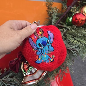May include: Red crocheted coin purse featuring an embroidered Stitch character decorated with Christmas lights. The purse has a silver clasp and is displayed with Christmas ornaments and greenery.