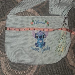 Puede incluir: Un bolso de hombro de crochet azul claro con un personaje azul Stitch de Lilo & Stitch. El bolso tiene un borla y el texto "Ohana means family" bordado en &eacute;l.