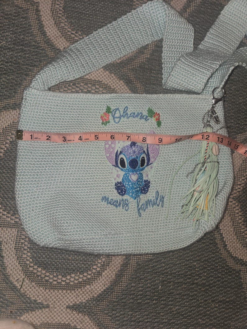 Puede incluir: Un bolso de hombro de crochet azul claro con un personaje azul Stitch de Lilo & Stitch. El bolso tiene un borla y el texto "Ohana means family" bordado en &eacute;l.