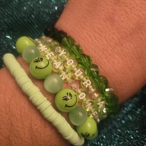 May include: A stack of bracelets in shades of green. One bracelet features the phrase "YOU'RE A MEAN ONE" spelled out with white letter beads. Other bracelets include smiley face beads and round beads.