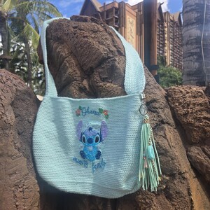 Puede incluir: Una bolsa de crochet azul claro con un personaje de Stitch bordado en la parte delantera. El bordado dice "Ohana means family". La bolsa tiene un borla azul claro.