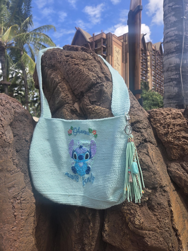 Puede incluir: Una bolsa de crochet azul claro con un personaje de Stitch bordado en la parte delantera. El bordado dice "Ohana means family". La bolsa tiene un borla azul claro.