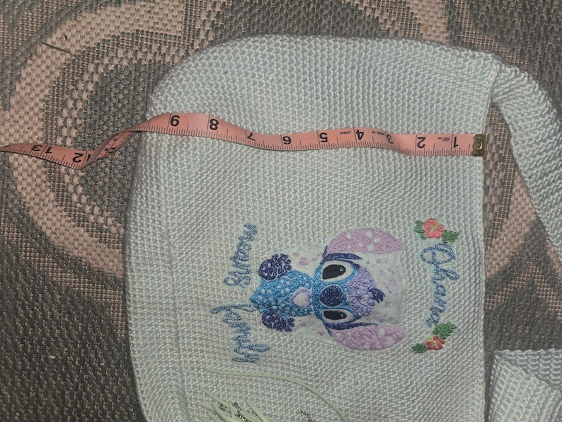 Puede incluir: Bolso blanco de crochet con un personaje de Stitch bordado en azul y morado, de la pel&iacute;cula de Disney Lilo & Stitch. El bordado incluye el texto "Ohana means family".