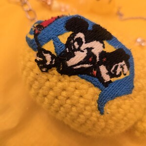 May include: A yellow, crocheted keychain with a blue and yellow patch featuring a black and white cartoon character. The keychain has a silver chain and small beads. The background is a solid yellow.