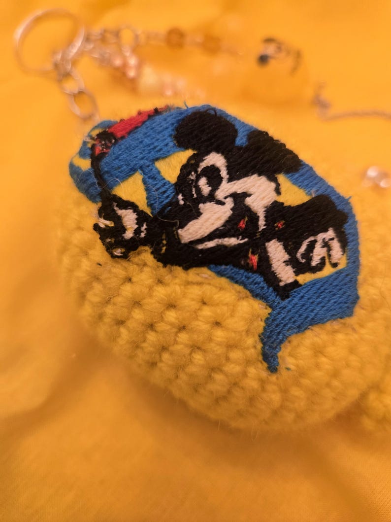 May include: A yellow, crocheted keychain with a blue and yellow patch featuring a black and white cartoon character. The keychain has a silver chain and small beads. The background is a solid yellow.