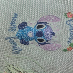 Puede incluir: Un dise&ntilde;o bordado en azul claro y morado de un personaje de Stitch de la pel&iacute;cula de Disney Lilo & Stitch. El dise&ntilde;o est&aacute; en una tela blanca con las palabras "My Family Always" y "Ohana" bordadas en azul.