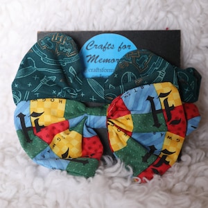 May include: A green and blue fabric bow with a Hogwarts house pattern. The bow is tied with a yellow, red, and green fabric. The bow is on a white fluffy surface. The text "Crafts for Memories" is visible on a black background.