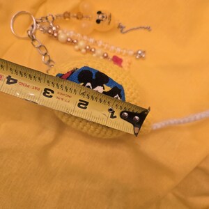 May include: A yellow crocheted keychain with a Mickey Mouse design, measuring approximately 1.5 inches in diameter. The keychain features a silver chain, beads, and a Mickey Mouse-shaped charm. The background is a solid yellow color.