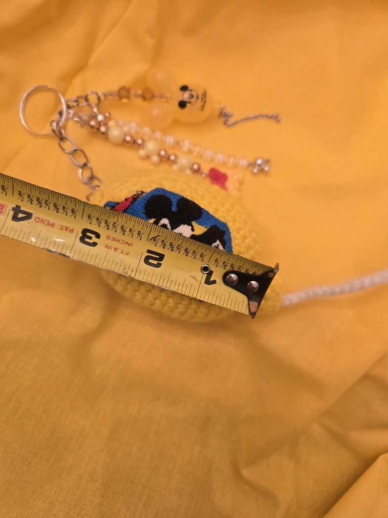 May include: A yellow crocheted keychain with a Mickey Mouse design, measuring approximately 1.5 inches in diameter. The keychain features a silver chain, beads, and a Mickey Mouse-shaped charm. The background is a solid yellow color.