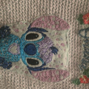 Puede incluir: Personaje de Stitch bordado en azul y morado con un fondo rosa. El texto "Ohana means family" est&aacute; bordado en azul en el lado derecho de la imagen.
