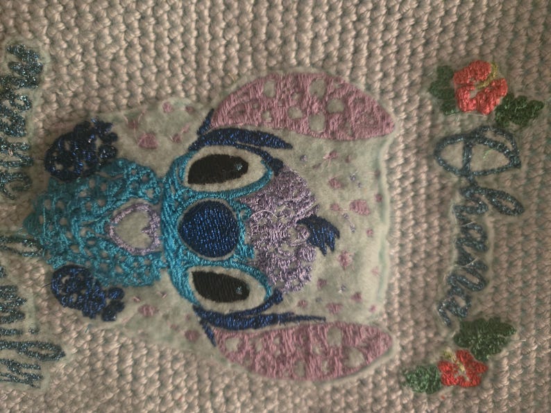 Puede incluir: Personaje de Stitch bordado en azul y morado con un fondo rosa. El texto "Ohana means family" est&aacute; bordado en azul en el lado derecho de la imagen.