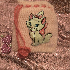 May include: A pink crocheted drawstring bag with a white embroidered cat design. The bag is sitting on a pink sparkly surface.