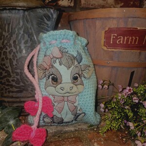 May include: A light blue crocheted drawstring bag with a pink ribbon and a pink heart design. The bag features an embroidered image of a brown and white cow with a pink bow. The bag is sitting on a wooden surface with a wooden bucket and green plants in the background.