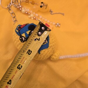 May include: A yellow crocheted keychain with a Mickey Mouse design, measuring approximately 4 inches in length. The keychain features a metal ring, beaded accents, and a white string. The background is a solid yellow.
