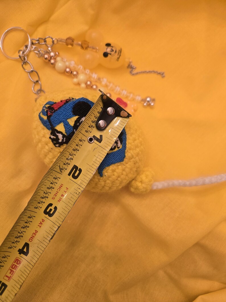 May include: A yellow crocheted keychain with a Mickey Mouse design, measuring approximately 4 inches in length. The keychain features a metal ring, beaded accents, and a white string. The background is a solid yellow.
