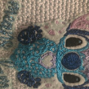 Puede incluir: Personaje Stitch bordado en azul y morado sobre un fondo de tela azul claro y blanco. El personaje tiene dos ojos negros grandes y un cuerpo con estampado azul y morado.