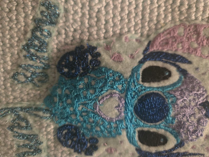Puede incluir: Personaje Stitch bordado en azul y morado sobre un fondo de tela azul claro y blanco. El personaje tiene dos ojos negros grandes y un cuerpo con estampado azul y morado.