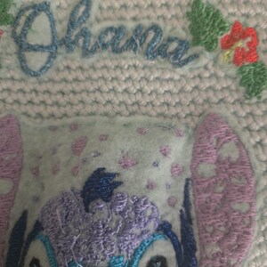Puede incluir: Personaje bordado en azul y morado con la palabra "Ohana" en bordado azul sobre un fondo azul claro.