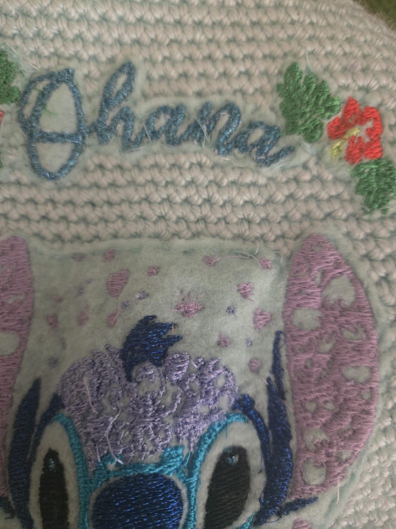 Puede incluir: Personaje bordado en azul y morado con la palabra "Ohana" en bordado azul sobre un fondo azul claro.