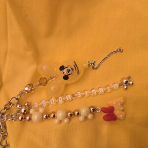 May include: A Mickey Mouse keychain with a clear, yellow Mickey Mouse head charm and a beaded charm spelling out "MINNIE MOUSE". The keychain also features a red and yellow bow charm and a silver chain.