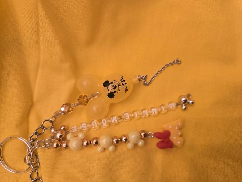 May include: A Mickey Mouse keychain with a clear, yellow Mickey Mouse head charm and a beaded charm spelling out "MINNIE MOUSE". The keychain also features a red and yellow bow charm and a silver chain.