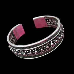May include: A silver cuff bracelet with an open design. The bracelet features intricate filigree and beaded details. The interior of the bracelet is a pinkish-red color. The bracelet is set against a black background.