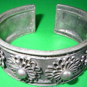 Puede incluir: Brazalete de puño plateado con diseño floral. El brazalete presenta una serie de flores estilizadas y detalles redondeados. El diseño abierto facilita su uso. El brazalete se muestra sobre un fondo verde.