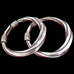 May include: Two silver bangles with a twisted design. Each bangle has coiled bands at the ends. The bangles are set against a black background, highlighting their metallic sheen and intricate details.