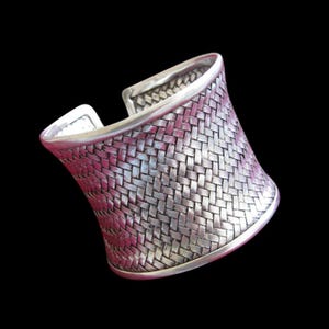 May include: Silver-toned cuff bracelet with a woven basket weave design. The wide bracelet has a curved shape and is set against a black background. The cuff is made of metal.
