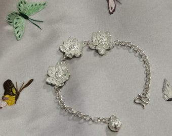 May include: A silver bracelet featuring three flower charms with clear gemstone centers. The bracelet has a delicate chain and a clasp closure. The background is a gray fabric with embroidered butterflies in various colors.