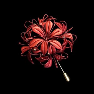 May include: A red floral lapel pin with a gold-colored pin backing. The pin features a detailed, three-dimensional design of a flower with multiple petals and small, silver-colored accents. The background is solid black.