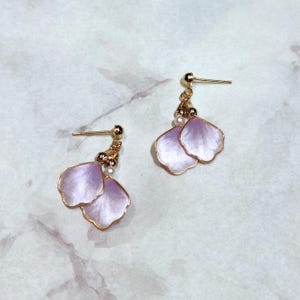 May include: A pair of stud earrings featuring gold-tone metal posts and small gold beads. Each earring displays two delicate, light purple, petal-shaped charms with a pearlescent finish and gold-coloured edges, suspended from the post.