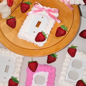 May include: Whimsical light switch and outlet covers featuring a strawberry and whipped cream design. The covers are white and pink, decorated with red strawberries and a creamy texture. One cover is accented with a pink ribbon.
