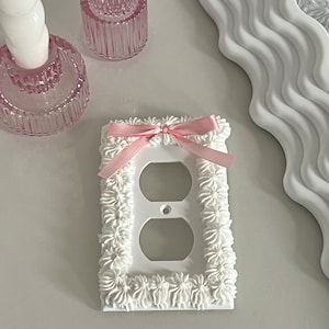Puede incluir: Tapa de tomacorriente blanca con un lazo rosa y un borde decorativo blanco. El borde está hecho para parecerse a la cobertura.