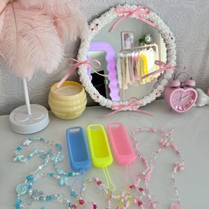 Może przedstawiać: Dekoracyjna aranżacja z okrągłym lustrem z białym kwiatowym obramowaniem i różowymi kokardkami. Obecne są również koralikowe zawieszki do telefonu oraz trzy kolorowe etui na telefon w kolorze niebieskim, żółtym i różowym.