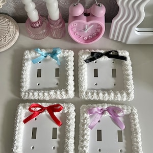 May include: Four white light switch covers with decorative frosting-like trim. Each cover features a different colored satin ribbon bow: light blue, black, red, and lavender.
