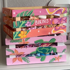 Puede incluir: Tres cajas de madera rosa con un tema tropical. La caja superior tiene la palabra "Bikinis" pintada en ella. La caja del medio tiene una tortuga verde y amarilla pintada en ella. La caja inferior tiene una flor rosa pintada en ella.