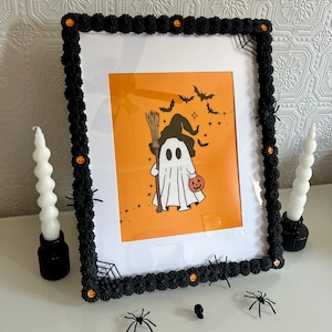 Könnte beinhalten: Ein schwarzer Bilderrahmen mit einem weißen Passepartout und orangefarbenem Hintergrund. Der Rahmen ist mit schwarzen und orangefarbenen Halloween-Verzierungen verziert. Das Bild im Rahmen ist ein Cartoon-Geist, der einen Hexenhut trägt und einen Kürbis hält.