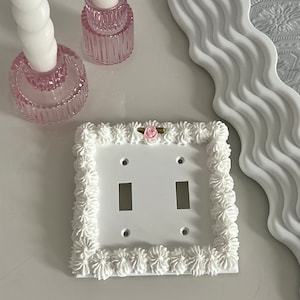 Op de afbeelding: Een witte dubbele lichtschakelaar met een decoratieve witte frosting rand en een roze roos accent.