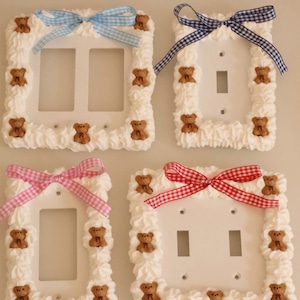 May include: Four white light switch covers decorated with white frosting-like trim and small brown teddy bears. Each cover has a different colored bow: blue, navy, pink, and red gingham. A charming, whimsical design.