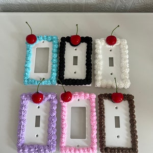 Puede incluir: Seis tapas de interruptor de luz con forma de pasteles con glaseado y una cereza en la parte superior. Los pasteles son de diferentes colores: azul, negro, blanco, morado, rosa y marrón.