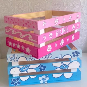 Puede incluir: Tres cajas de almacenamiento de madera apiladas una encima de la otra. La caja superior está pintada de rosa con conchas blancas y la palabra "Bikinis". La caja del medio está pintada de rosa con estrellas blancas. La caja inferior está pintada de azul con flores de hibisco blancas.