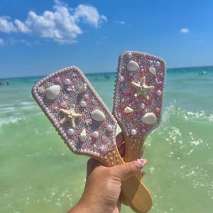 May include: Two decorative hairbrushes with wooden handles are held up against a backdrop of the ocean and sky. The brushes are embellished with pink and white beads, seashells, and starfish. The handles are made of light-colored wood.
