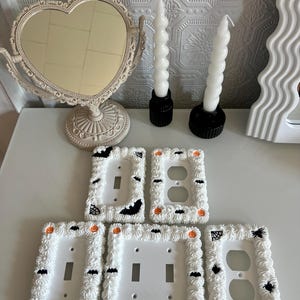 May include: Five white light switch covers decorated with a Halloween theme. The covers feature white frosting, black bats, orange pumpkins, and black spiderwebs.