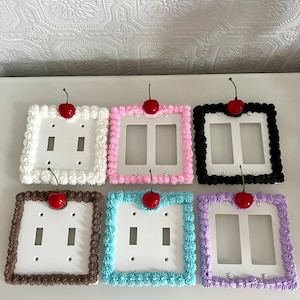 May include: Six decorative light switch covers with a cake frosting design and a red cherry on top. The covers are in various colors, including white, pink, black, brown, blue, and purple.